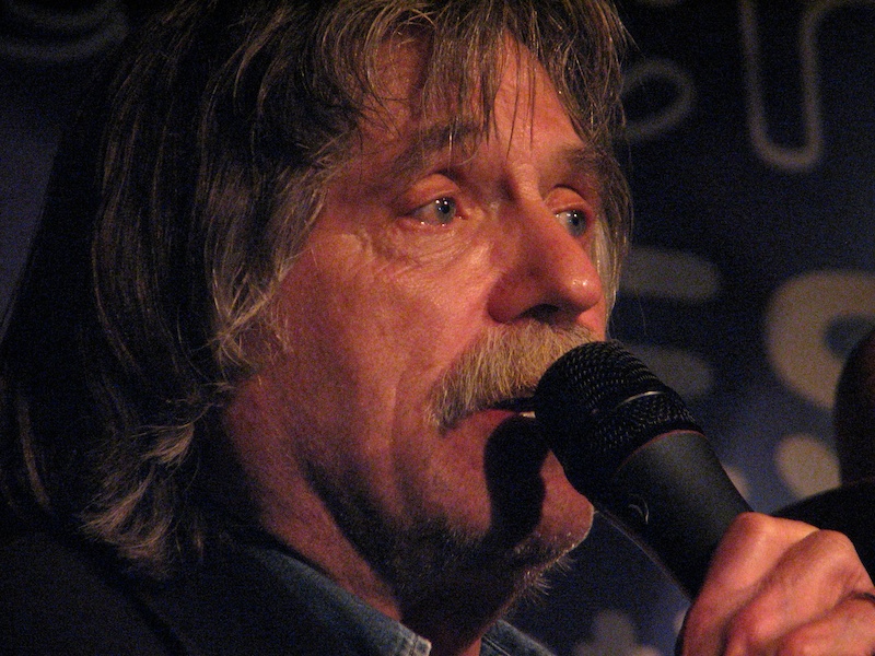A close-up photo of Johan Derksen’s face. He is standing in a dimly lit area, looking to the right and holding a microphone that he’s speaking into.