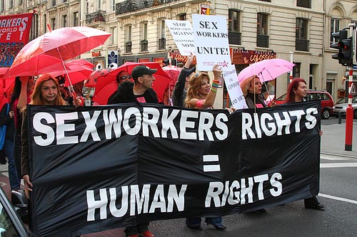Five people walking at the front of a march. They are holding a large black banner which says “sex workers rights equals human rights” in bold white text. One person is holding a white sign that says “Sex workers unite!”. People behind them are holding red umbrellas.